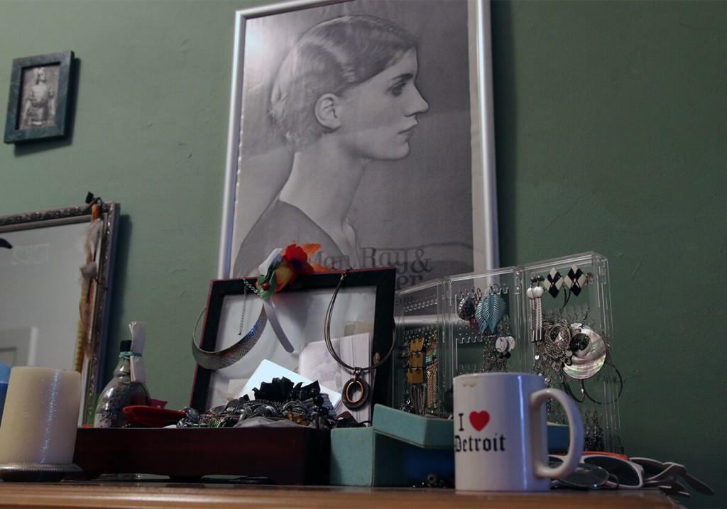 The top of a dresser that is covered with jewelry, an I Love Detroit coffee mug, pairs of glasses, and a candle; behind it is a Man Ray poster.